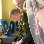 Groom signing the marriage certificate while bride and minister watch.